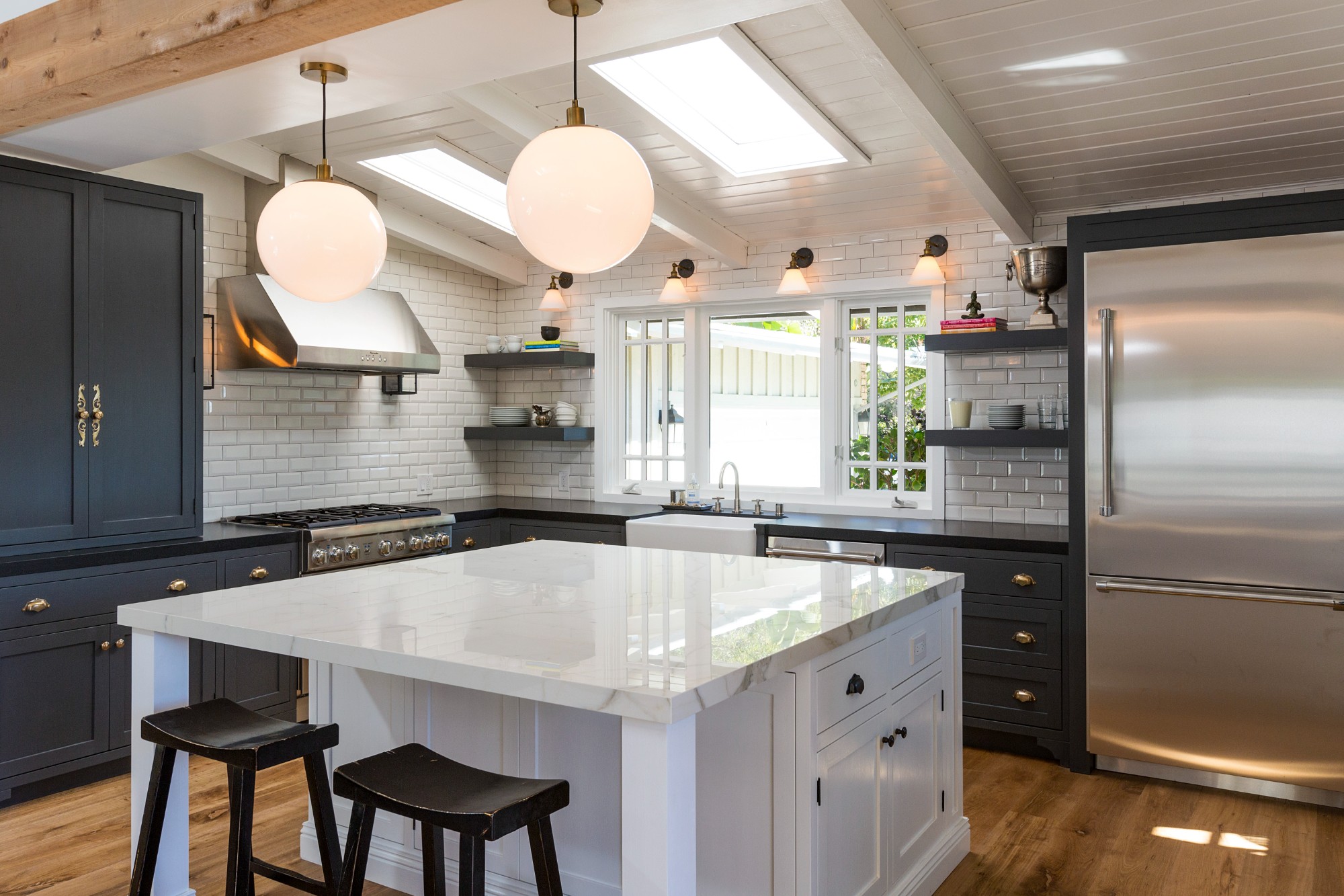 Beach house kitchen alternate angle, apron-front farmhouse sink, bridge faucet, brass cup pulls, dual skylights overhead, white globe pendants. Pacific Palisades.