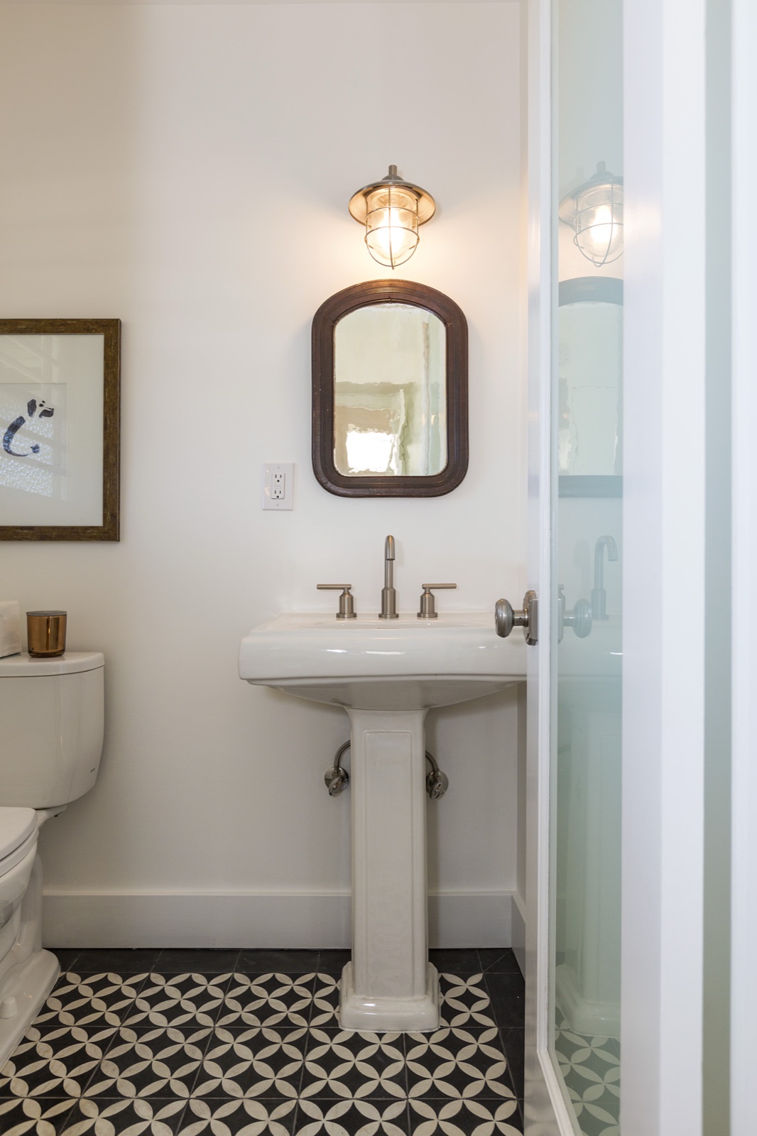 Powder room at 855 Jacon Way Pacific Palisades, vintage white pedestal sink with widespread satin-nickel faucet, arched apothecary mirror in a dark frame, caged wall sconce overhead, patterned black-and-white cement-tile floor. Coastal cottage powder room by ESRL Development.